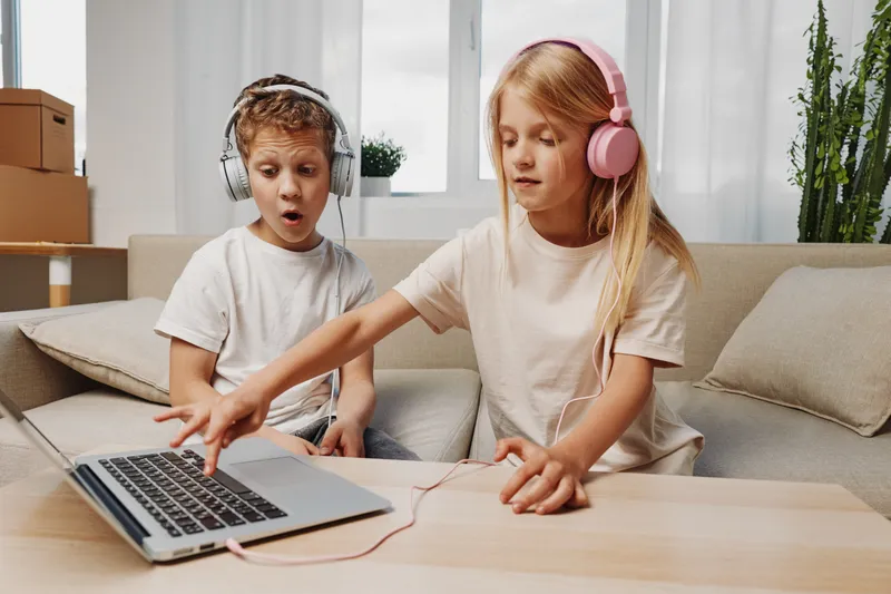 Two children with headphones use a laptop on a wooden table in a cozy living room, showing curiosity and focus while engaging with digital content and technology together.