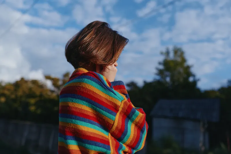 Woman in a colorful striped sweater with short hair outdoors, looking down in a contemplative pose. Casual outfit, autumn light, cozy knitted jumper and soft blue sky background.