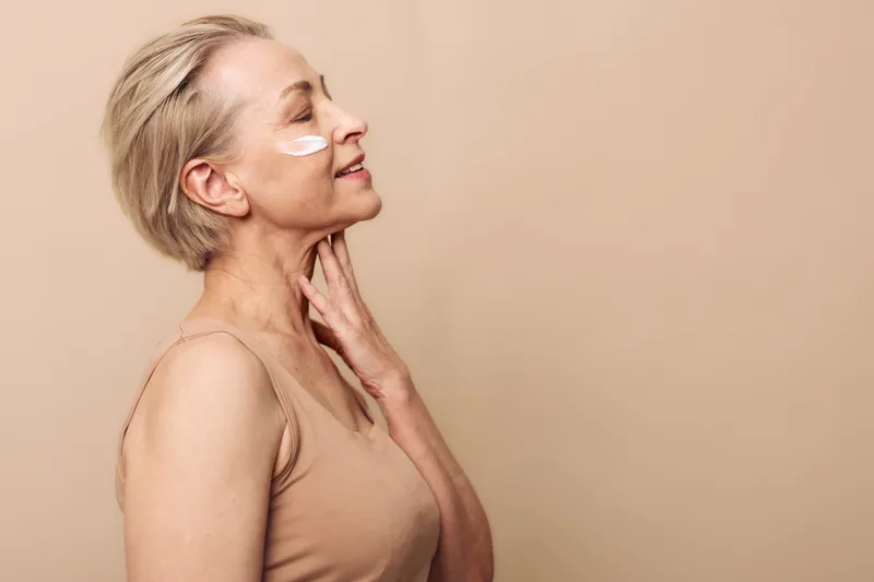 Smiling mature woman with short blonde hair applies white cream under eye in beige tank top. Side profile on soft beige background. Concept of skincare, beauty, antiaging, and selfcare.