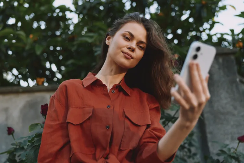 Young woman in a rust red shirt takes a casual selfie in a sunny garden, smiling softly as she angles the camera