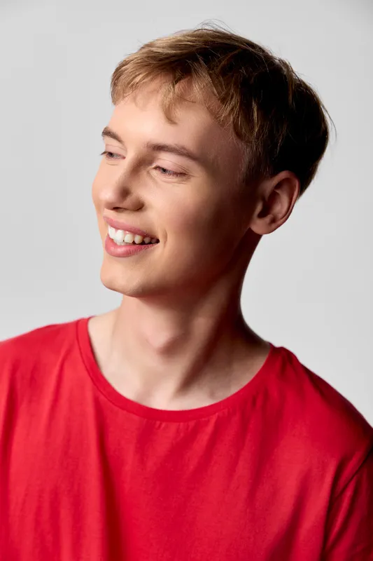 Smiling man in a red shirt looking to the side, with a warm expression and relaxed posture, studio lighting highlighting cheerful mood, casual fashion, candid moment, clear focus on smile and