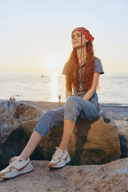 young woman sitting on rocks by the sea during sunset wearing casual denim outfit and red bandana with relaxed and calm mood in warm natural light