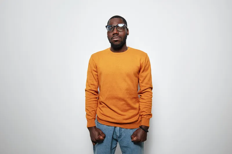 African American man with dark skin wearing orange sweater and glasses standing against white background looking confident and serious with clenched fists and casual jeans.