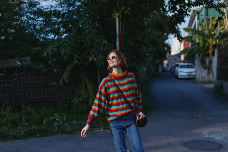 Woman in a colorful striped sweater and sunglasses smiling while walking in jeans with a crossbody bag on a quiet suburban street at evening, casual travel lifestyle vibe.
