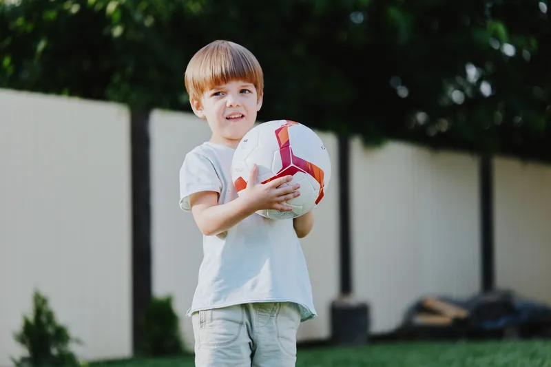 Happy boy holding a soccer ball, showcasing his enthusiasm and love for sports in a sunny outdoor setting with lush green grass and a blurred background.