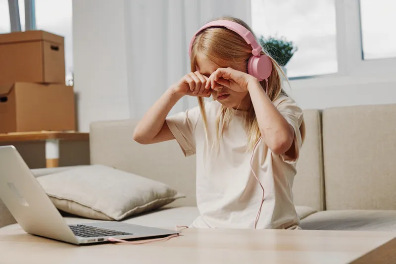 Tired little girl in headphones rubs eyes sitting on couch with laptop, feeling sleepy and distracted during online learning at home.