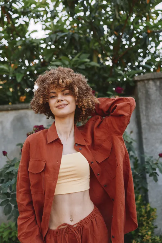 A stylish woman in a rust jacket and peach top poses outdoors, exuding confident casual lifestyle. Bright garden backdrop and natural light create a vibrant fashion portrait