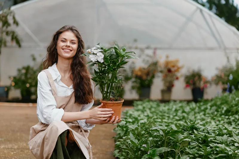Smiling young woman holding a potted plant in a greenhouse, showcasing vibrant greenery and a joyful gardening atmosphere