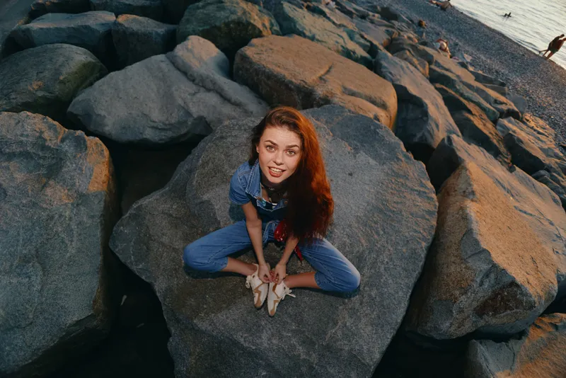 Lifestyle woman in modern boho-western denim street style sitting on rocks by the shore, with warm film color tones enhancing the natural outdoor vibe.