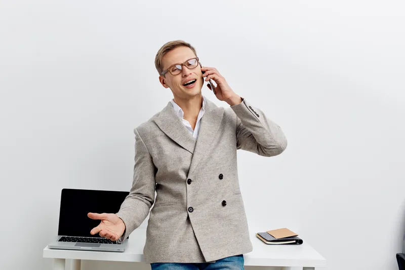 Smiling confident man wearing gray checked blazer talking on mobile phone while standing at desk with laptop and notebook, casual work environment, professional business attire, joyful expression, off