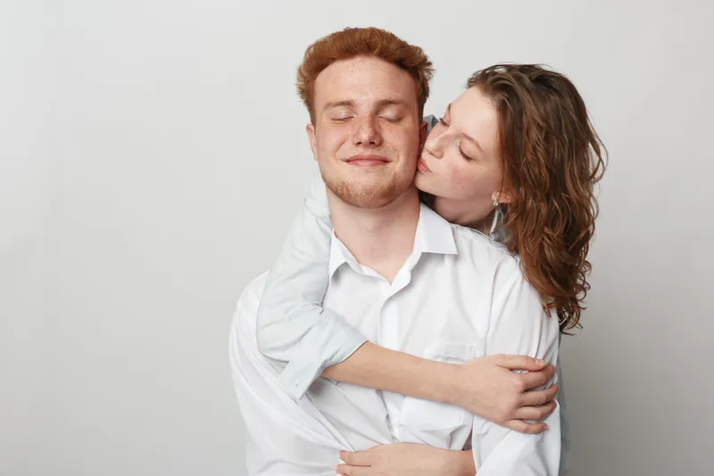 Young couple in white shirts embraces with eyes closed, woman kissing man on cheek. Intimate moment of love, affection, and tenderness on neutral background.