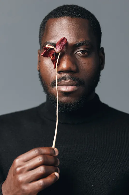 African man with dark skin holding dried flower in front of face in creative conceptual visual style. Portrait with black turtleneck and neutral background for artistic expression.