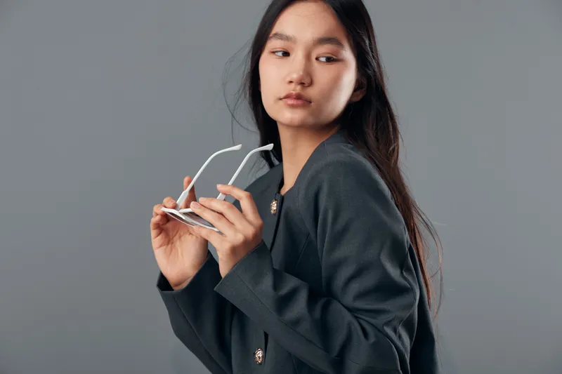 Young woman holding sunglasses with long hair looking aside in studio. Female portrait with casual dark jacket against gray background, thoughtful expression and natural makeup indoors.