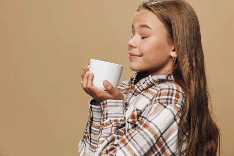 Girl in plaid shirt holding white cup close to face with closed eyes enjoying warm beverage on beige background. Relaxation, comfort and cozy lifestyle for youth satisfaction.
