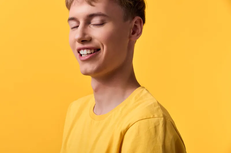 Joyful male model in a yellow shirt with a closed eyes smile against a vivid yellow background, conveying warmth, positivity, and vibrant lifestyle appeal for fashion or wellness visuals