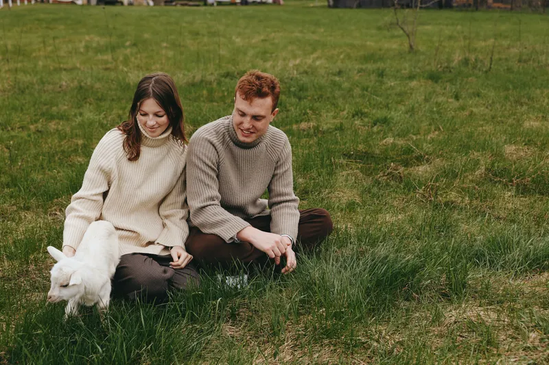 Young couple in cozy sweaters sits on green grass in a park playing with a white baby goat enjoying a relaxed lifestyle moment on a cloudy day outdoors in nature.