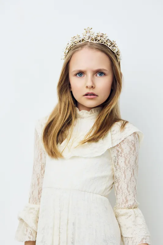 Young girl in white lace dress with crown, expressing elegance and innocence against a soft neutral background, perfect for childhood or fashion themes