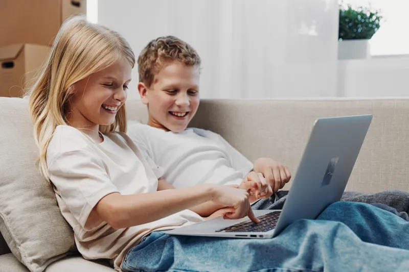 Two happy children sitting on a couch using a laptop together, smiling and enjoying digital learning or entertainment in a bright living room with natural light.