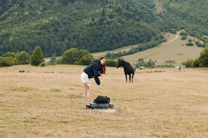 A woman in a rural field bends over a backpack while a horse grazes nearby, with rolling hills in the distance, creating a calm and natural outdoor scene.