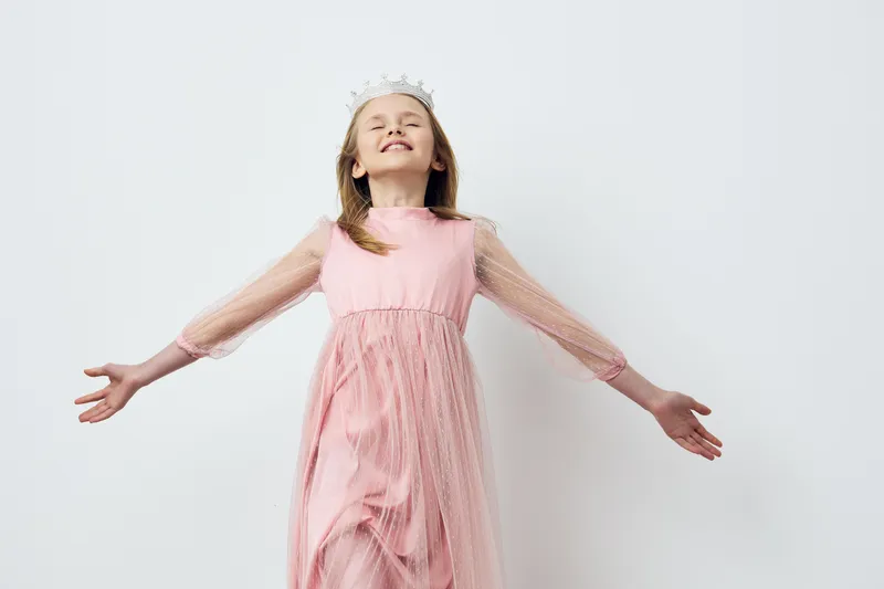 Young girl in a pink dress with sheer sleeves joyfully posing against a simple white background, exuding happiness and innocence