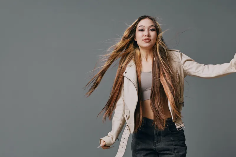 Young woman with long hair wearing beige jacket and black jeans poses confidently against gray background in studio for casual fashion and beauty portrait photography.