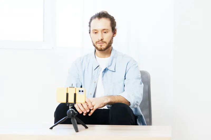 Young man filming a vlog indoors, wearing a casual blue shirt, expressing focus and creativity in a bright, minimalistic setting