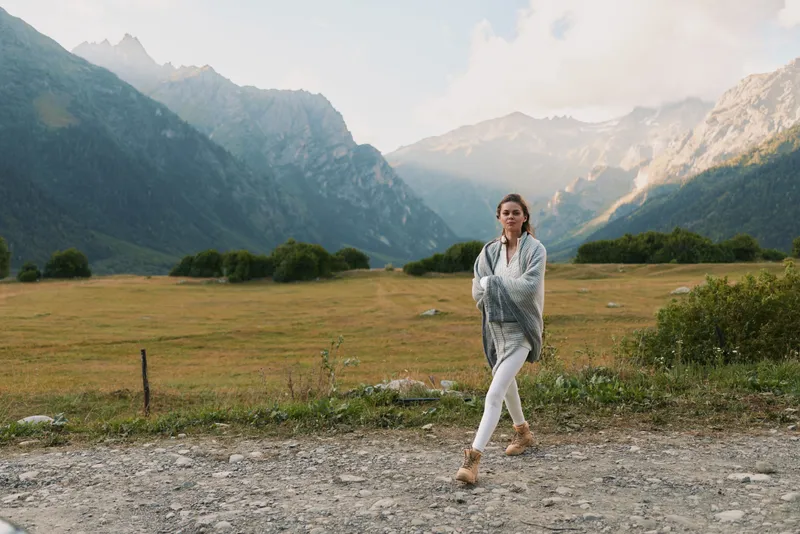 Young woman walks along a gravel road in a mountain valley, calm and thoughtful, framed by distant peaks, that suggest solitude, exploration, and a sense of open space.