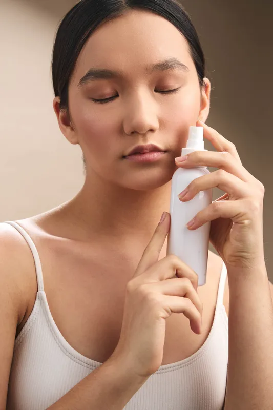 asian woman applies skincare spray to her face in a close up studio shot, calm and focused expression, soft lighting, minimal background creating a serene beauty scene.