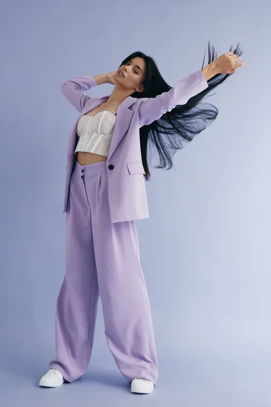 Young woman in lilac pantsuit and white lace top posing confidently with flowing hair against pastel background, showcasing modern fashion style and elegance.