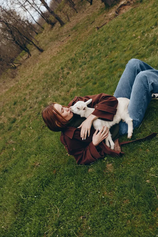 Woman lying on grass in a park, cuddling a white goat kid, enjoying a relaxed outdoor moment, soft light, natural colors, casual outfit, tranquil lifestyle scene