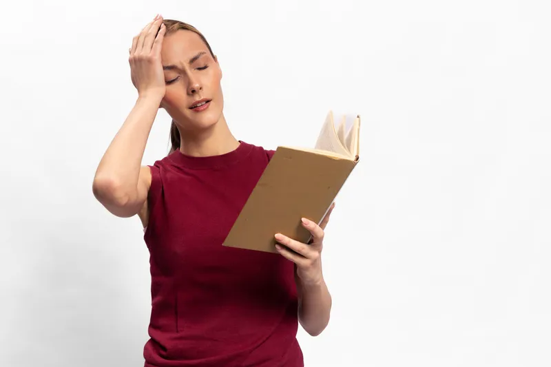 Young woman in red sleeveless top reads a book with a confused expression, hand on forehead. White background highlights her thoughtful and puzzled mood during study.