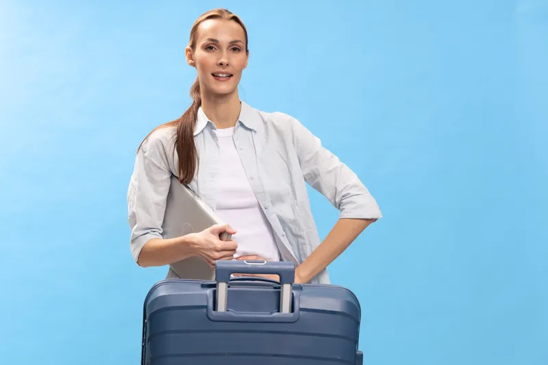 Young woman traveler with suitcase and laptop stands on blue background smiling confidently. Concept for business travel, vacation preparation, and modern lifestyle.