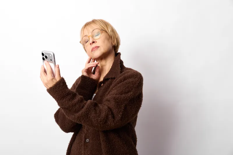 Senior woman with glasses in brown coat holds smartphone and answers calling, focused expression, communication concept, isolated on light background, indoors