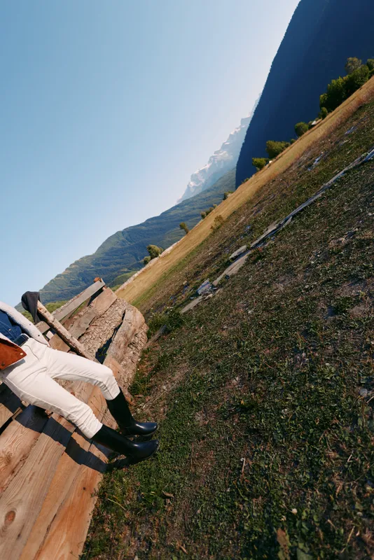 Rider in black boots and white breeches sits on a wooden bench in mountains meadow countryside outdoors, enjoying sunny alpine landscape and relaxed equestrian leisure moment.