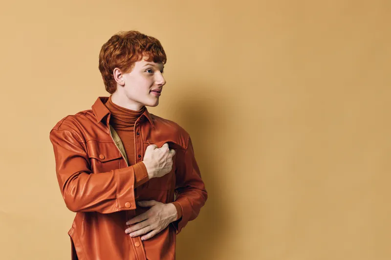 Young man with red hair wearing brown leather jacket and turtleneck standing and looking sideways on beige background in studio. Casual fashion and confident pose with space.