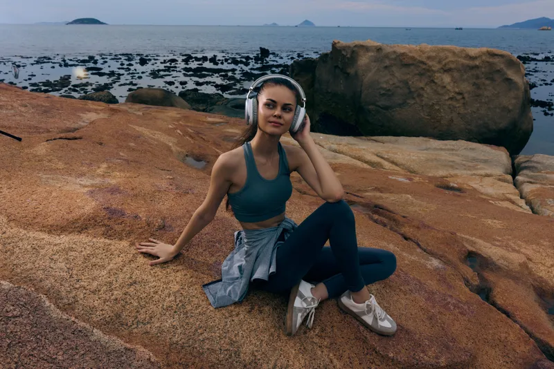 Fitness girl listening to music on rocky beach, wearing sporty outfit, enjoying tranquility and sunset, representing healthy lifestyle and active living