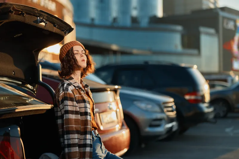Woman leaning on car trunk in urban parking, sunlight warming her face at golden hour, beanie and flannel outfit, candid lifestyle portrait conveying authenticity and mindful living.