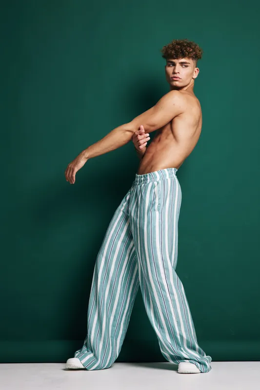 Young man posing shirtless in wide striped pants with curly hair in fashion studio. Male model demonstrating relaxed posture against plain green backdrop.