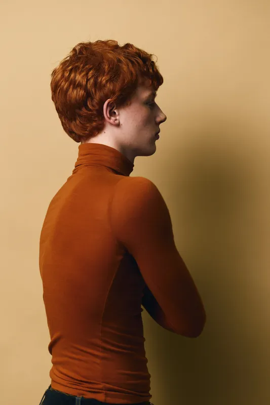 Profile of young redhead man standing with arms crossed wearing brown turtleneck against beige background in studio portrait with soft lighting.