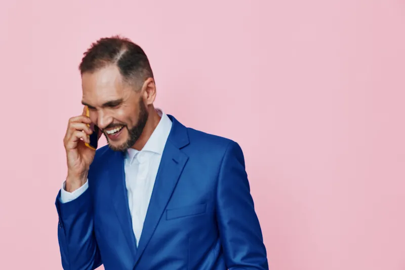 A professional man in a blue suit on a solid pink background, smiling while talking on the phone. This isolated colored backdrop highlights confident business style and positive emotion.