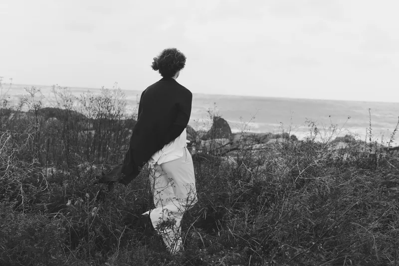 Silhouette of a woman walking through meadow near coast in grayscale, wind in hair, tranquil and solitary journey amid sea spray and wildflowers