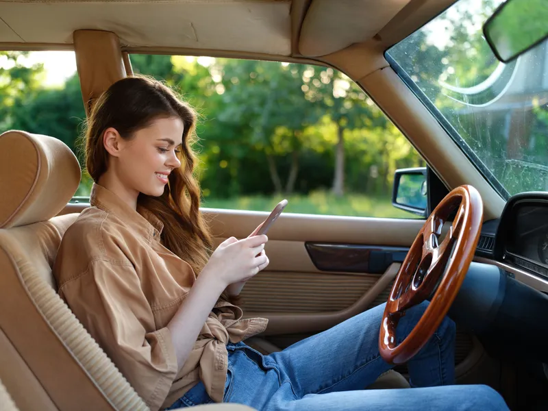 Young woman with long hair enjoying a peaceful moment in her stylish car, checking her phone while sitting comfortably in a lush green background.