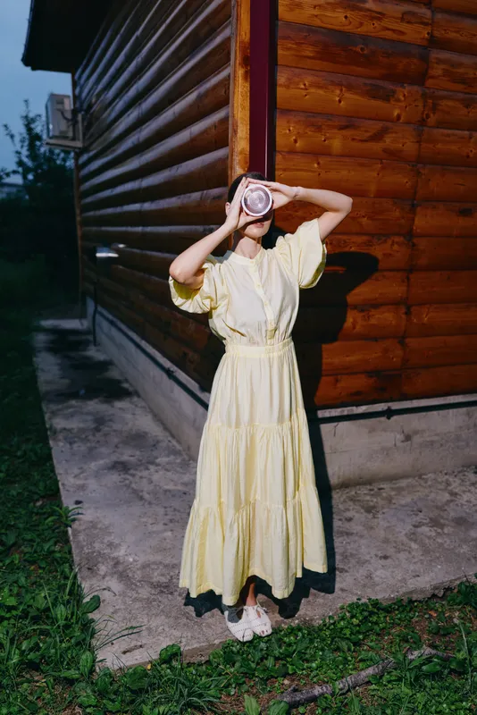 Woman in a yellow dress stands beside a wooden cabin at dusk, holding a camera to her face and taking a photo, creating a playful artistic moment.