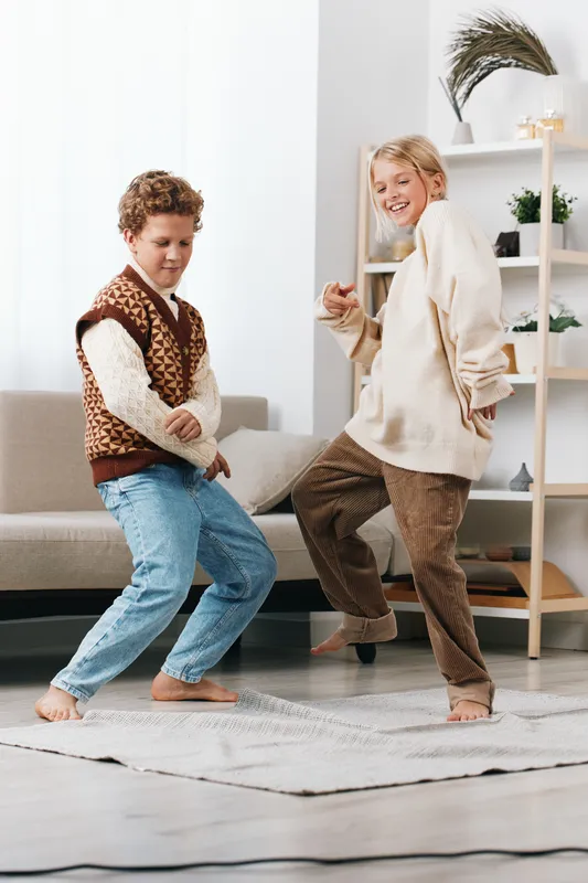 Two children dance playfully in a bright living room, wearing cozy sweaters and jeans, capturing lively movement, spontaneous smiles, and carefree energy.