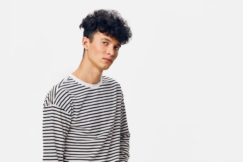 young man wearing black and white striped shirt with curly hair posing against white background, casual style and relaxed expression, studio portrait