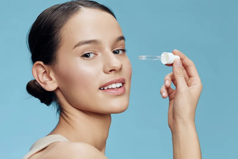 Young woman applying serum to her face, promoting healthy skincare routine with a joyful expression against a light blue background