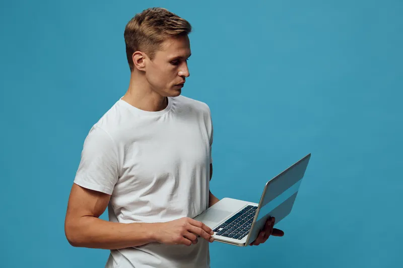 Young man in plain white t-shirt using laptop computer with a focused expression against a bright blue background. Studio portrait, technology, concentration, casual clothing, modern, isolated on blue