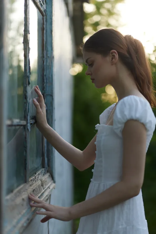 Thoughtful young woman in a white dress, gazing out of a vintage window, surrounded by nature, showcasing a serene and contemplative mood, perfect for lifestyle themes
