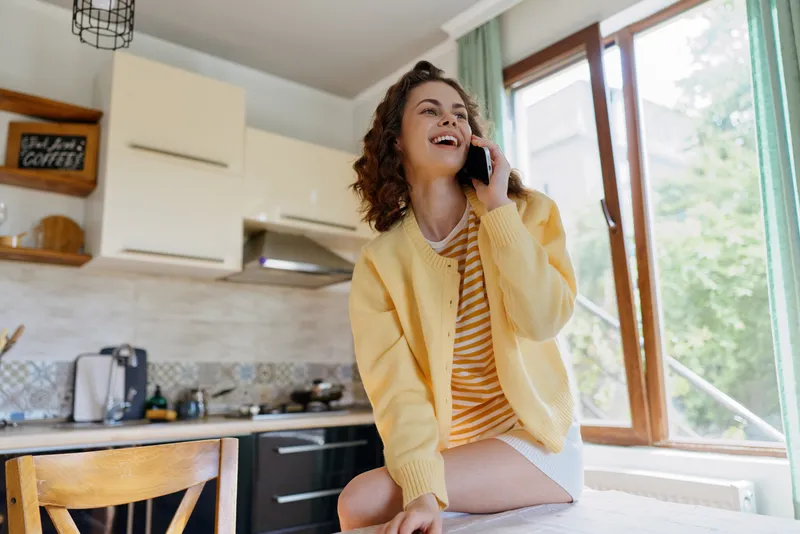 A smiling woman in a yellow cardigan sits in a bright kitchen, talking on a phone. A casual home lifestyle moment showing daily routine, comfort, and wellbeing.