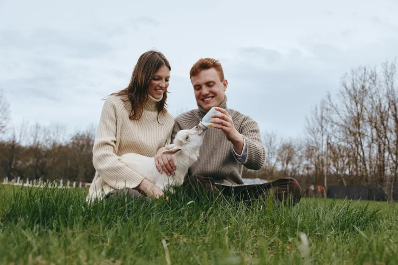 Joyful couple with a goat kid outdoors, sharing a warm moment on the grass. Cozy sweaters, natural light, rustic vibe. Lifestyle photography highlighting companionship, tenderness, and rural charm.